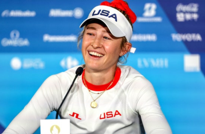 Nelly Korda in a white long-sleeved top and cap with USA printed on both smiles at a press conference. She sits in front of a blue background with various logos, speaking into a microphone.